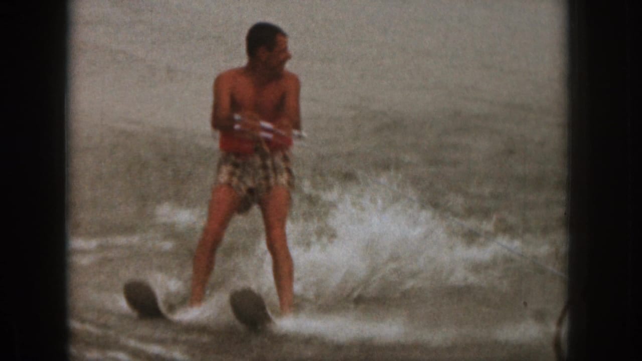 Man in red swim shirt and patterned trunks water skiing on a lake.