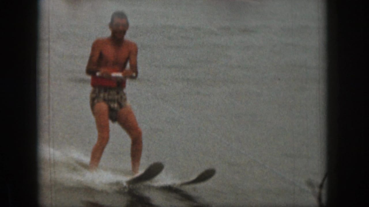In 1961 Michigan home-movie footage, a shirtless man in patterned trunks and a red life vest skis across a lake.