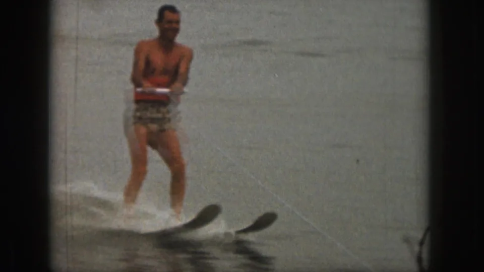 The skier smiles toward the camera in the 1961 Michigan lake footage, with twin skis visible near the water surface.