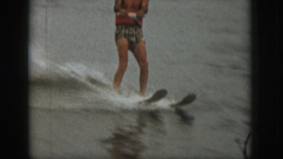 A closer, partly cropped 1961 Michigan frame shows the man's legs, tow handle, skis, and spray as he rides across the lake.