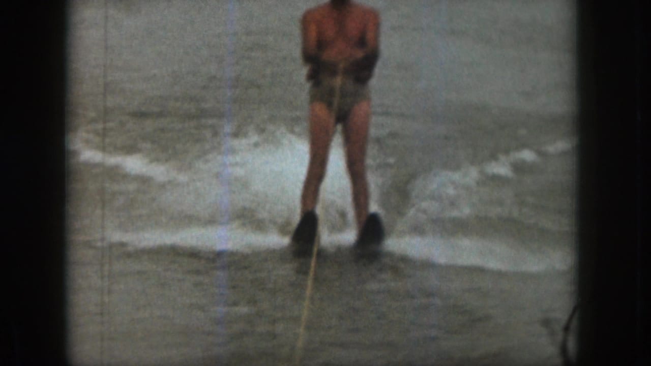 1961 Michigan home-movie frame showing an adult man in swim trunks water skiing on a lake, with tow rope and spray visible.