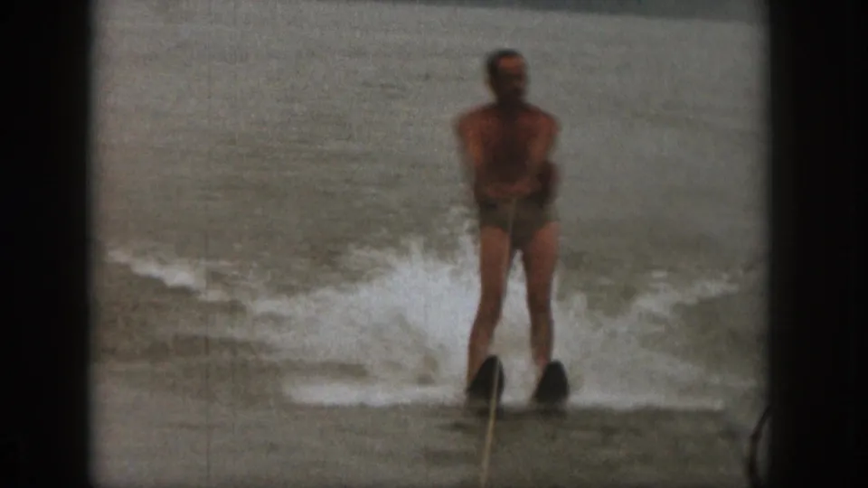 1961 Michigan lake footage frame with an adult man on water skis facing the camera while a wake breaks behind him.