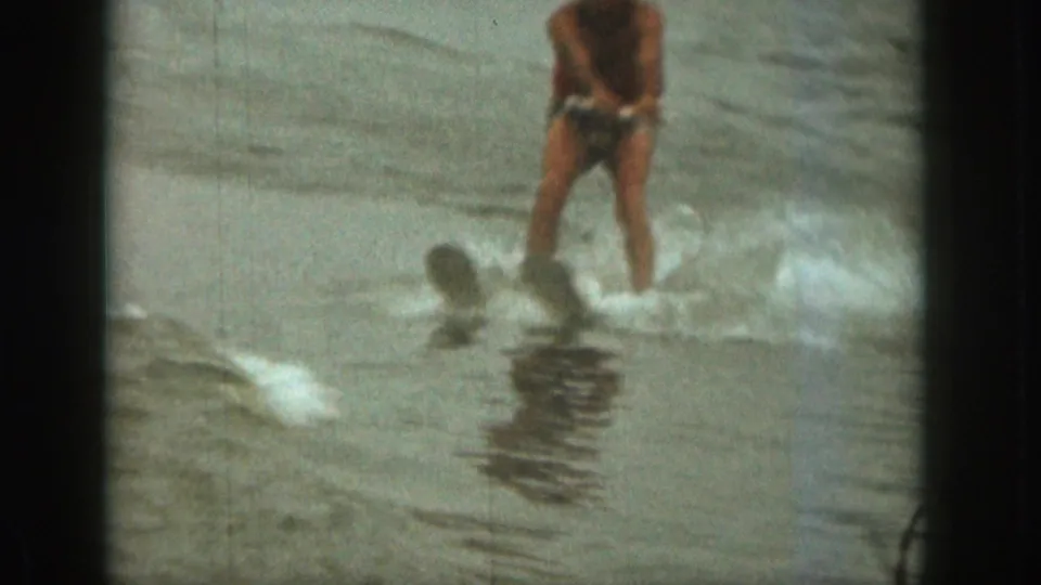1961 Michigan 8mm frame showing a shirtless adult water skier in swim trunks moving through spray on choppy water.