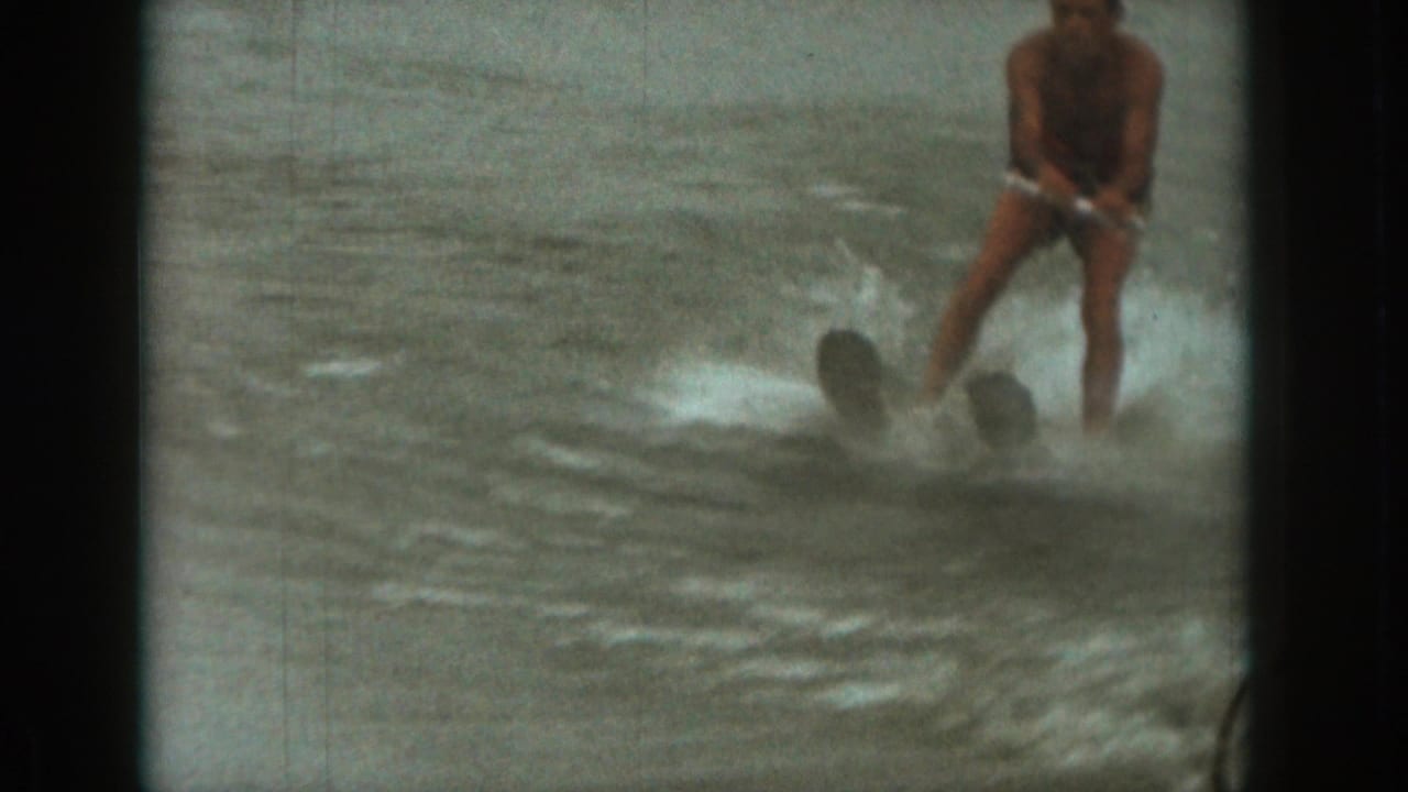 1961 Michigan 8mm frame showing a man water skiing on a lake, holding a tow handle as spray rises around him.