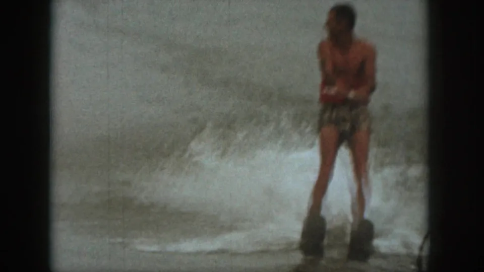 1961 Michigan home-movie frame of a man in a red life vest skiing upright over choppy lake water.
