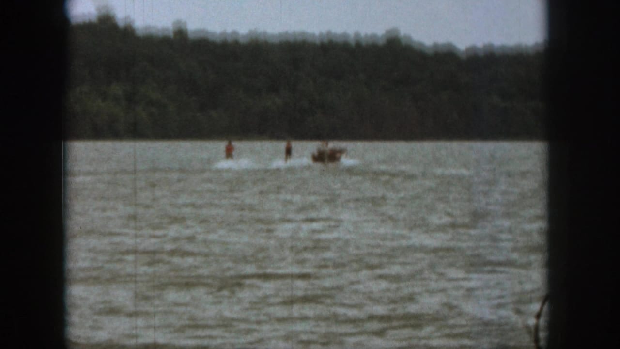 Distant water skier and small boat move across a lake near a wooded shore.