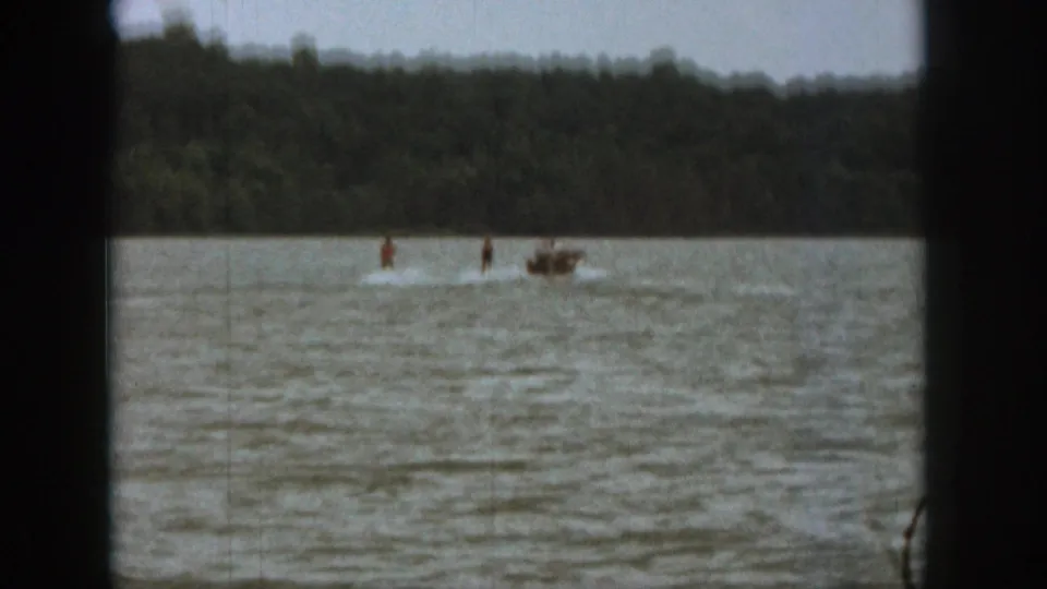 Wide 1961 Michigan lake view with a small motorboat pulling a distant water skier against a tree-lined shoreline.