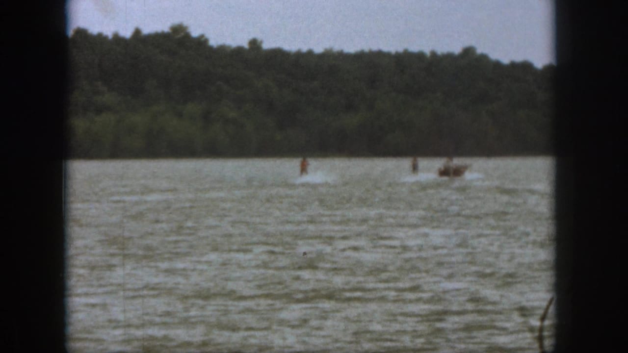 1961 Michigan home-movie frame with a distant water skier and motorboat crossing the lake below a wooded shoreline.