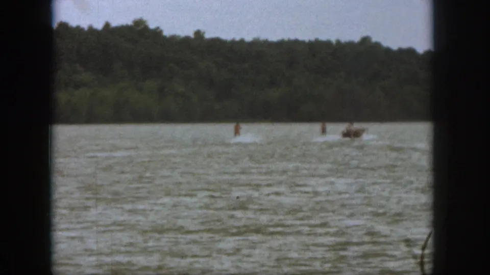 1961 Michigan home-movie frame with a distant water skier and motorboat crossing the lake below a wooded shoreline.