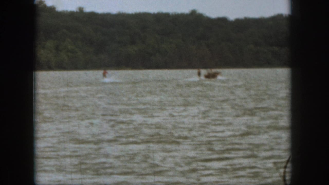 Wide 1961 Michigan lake frame showing a small motorboat and distant water skier moving along the tree-lined shore.