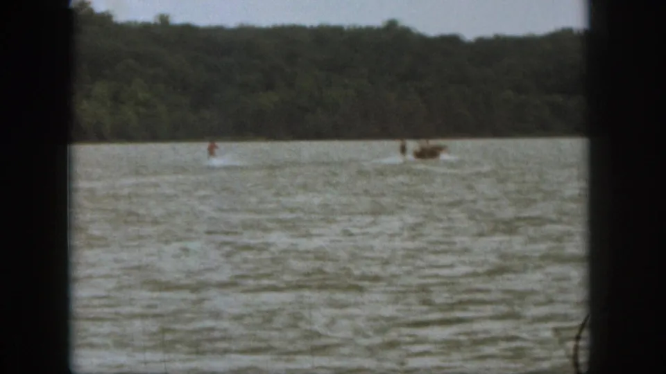 Wide 1961 Michigan lake frame showing a small motorboat and distant water skier moving along the tree-lined shore.