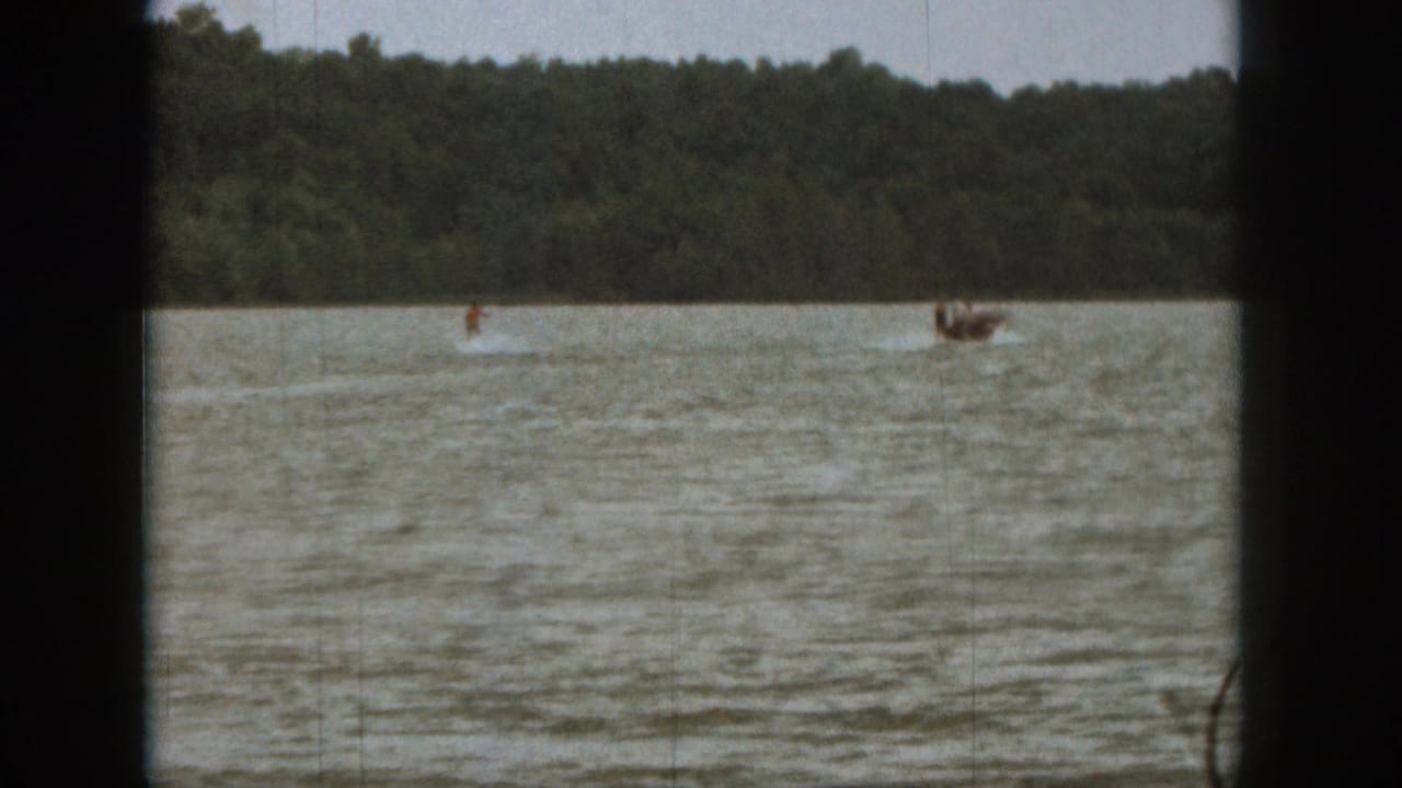 1961 Michigan 8mm frame showing a distant water skier and towing boat on rippled lake water below a forested shore.