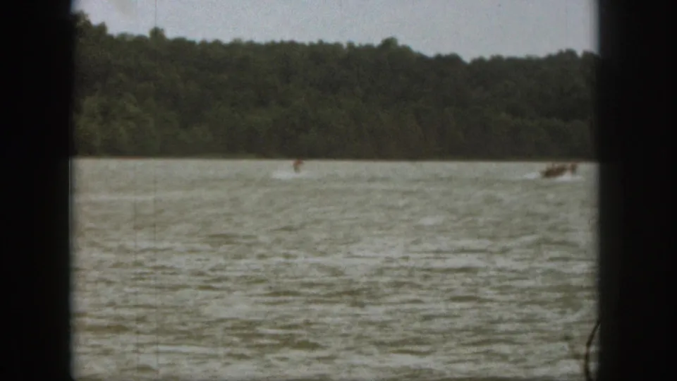 1961 Michigan home-movie frame with a distant water skier crossing choppy lake water near a towing motorboat.