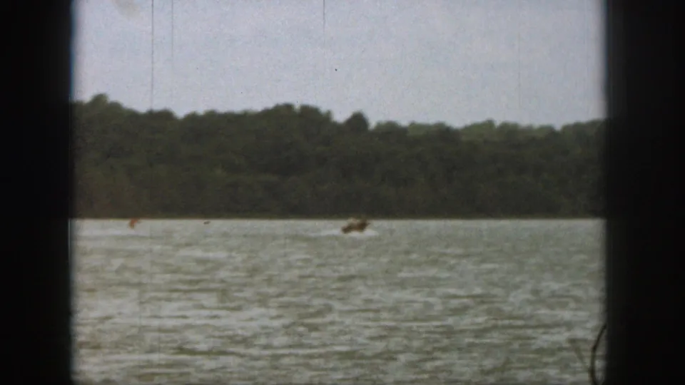 1961 Michigan home-movie frame showing a distant water skier crossing a lake near a forested shore.