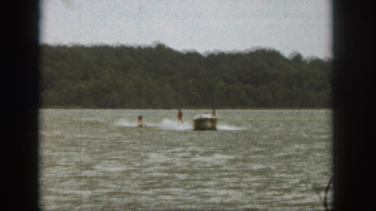 Motorboat pulls two water skiers across a lake near a wooded shore.