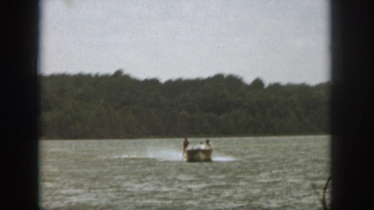 1961 Michigan home-movie frame showing a motorboat moving across a lake with a skier or rider near its wake.