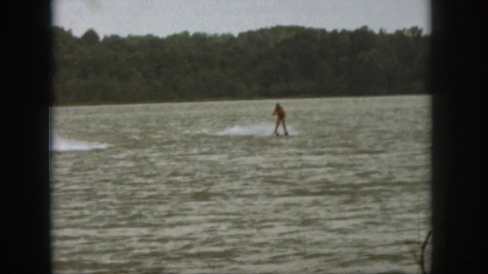 Distant rider in red swimwear moves across a Michigan lake in 1961 home-movie footage, with shoreline trees in the background.