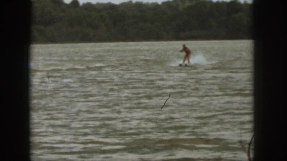 A distant water-sport rider is pulled across a Michigan lake, framed by 8mm borders and a wooded shoreline.