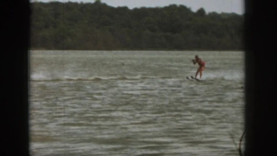 1961 Michigan home-movie frame of a towed rider in red swimwear crossing a lake, with spray and trees behind.