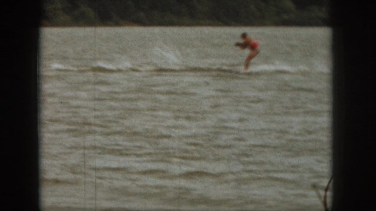 Board rider in red moves across open lake water near a low wooded shoreline.