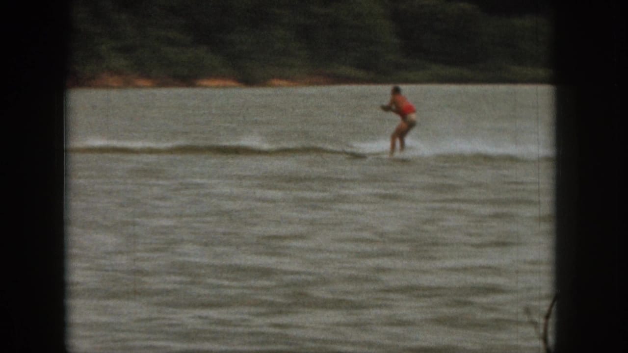 Michigan 1961 home-movie frame showing a distant board rider in red with wake spray on a calm lake.