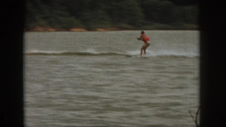 Michigan 1961 home-movie frame showing a distant board rider in red with wake spray on a calm lake.
