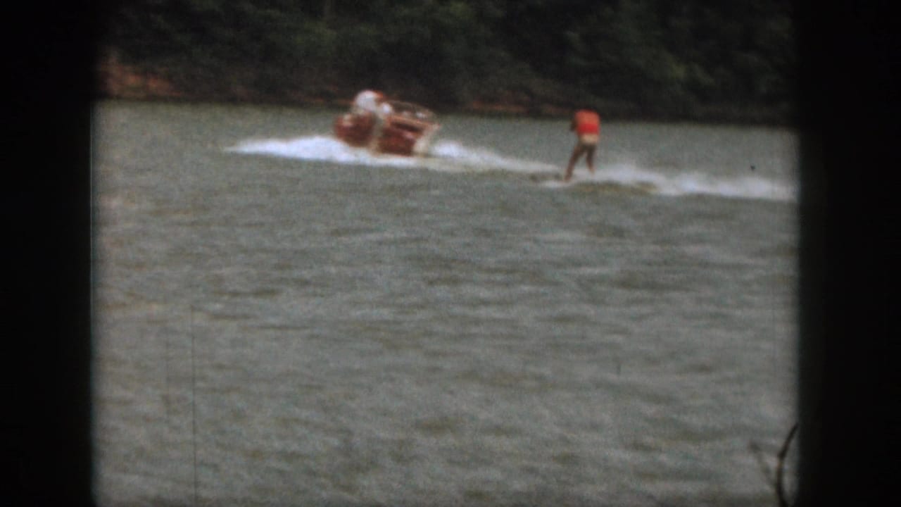 In 1961 Michigan home-movie footage, a motorboat pulls a water skier in a red top across a lake bordered by trees.