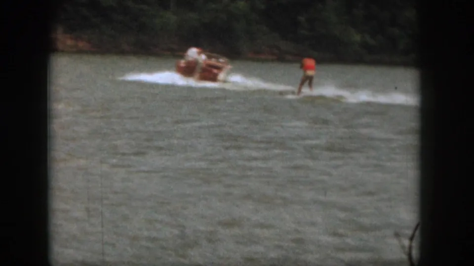 In 1961 Michigan home-movie footage, a motorboat pulls a water skier in a red top across a lake bordered by trees.