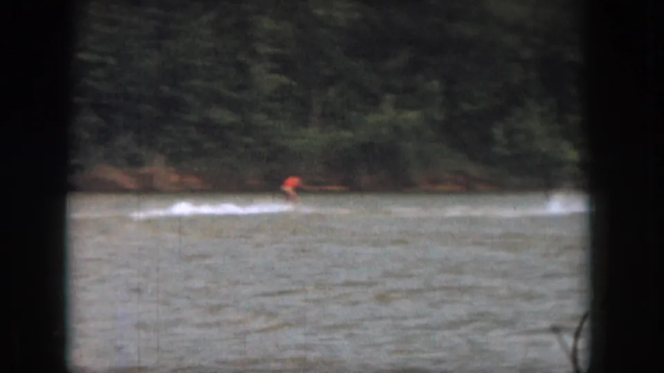 A water skier remains visible at a distance as wake trails across a wooded Michigan lake in 1961 8mm footage.