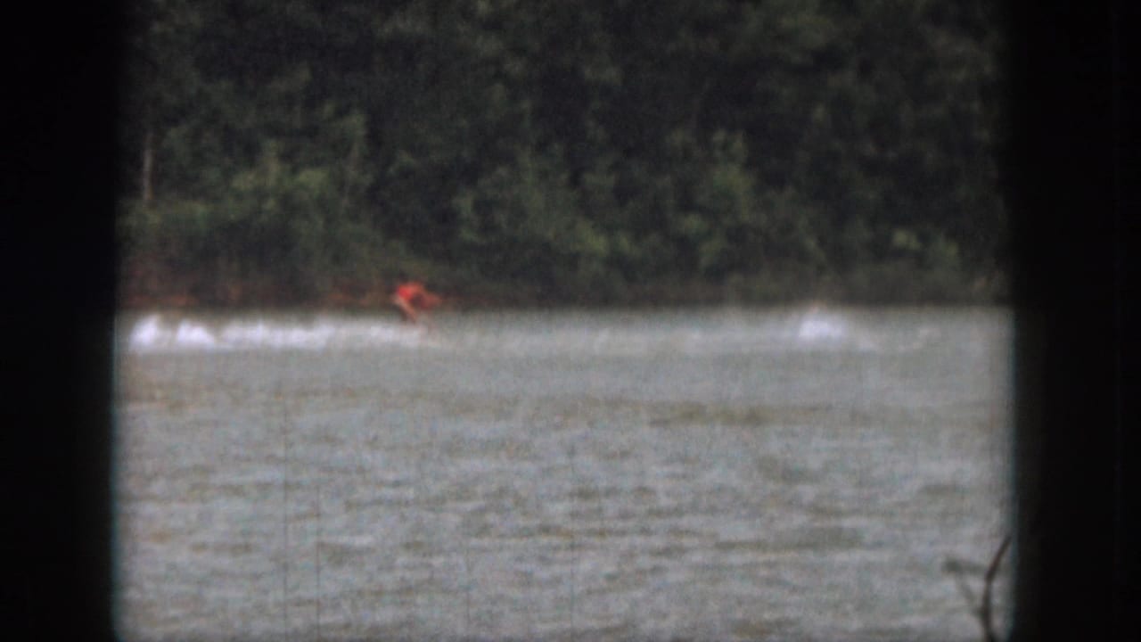 1961 Michigan 8mm frame showing a distant water skier in red crossing a lake with trees along the far shore.