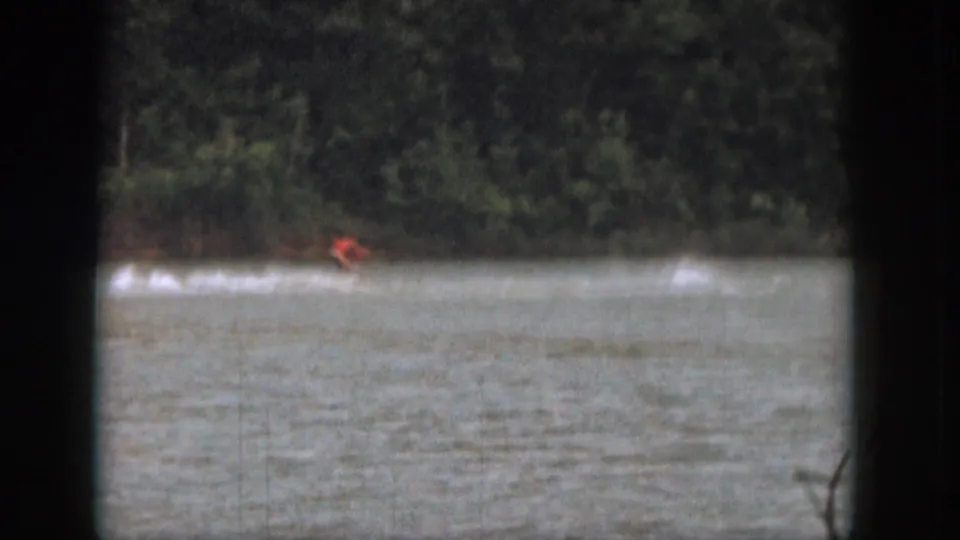 1961 Michigan 8mm frame showing a distant water skier in red crossing a lake with trees along the far shore.