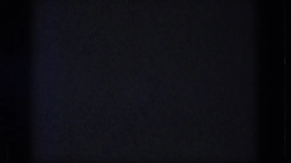 Dark 8mm home-movie frame with grain, low exposure, a small blue flare, and vertical film-edge shadows before the lake scene appears.