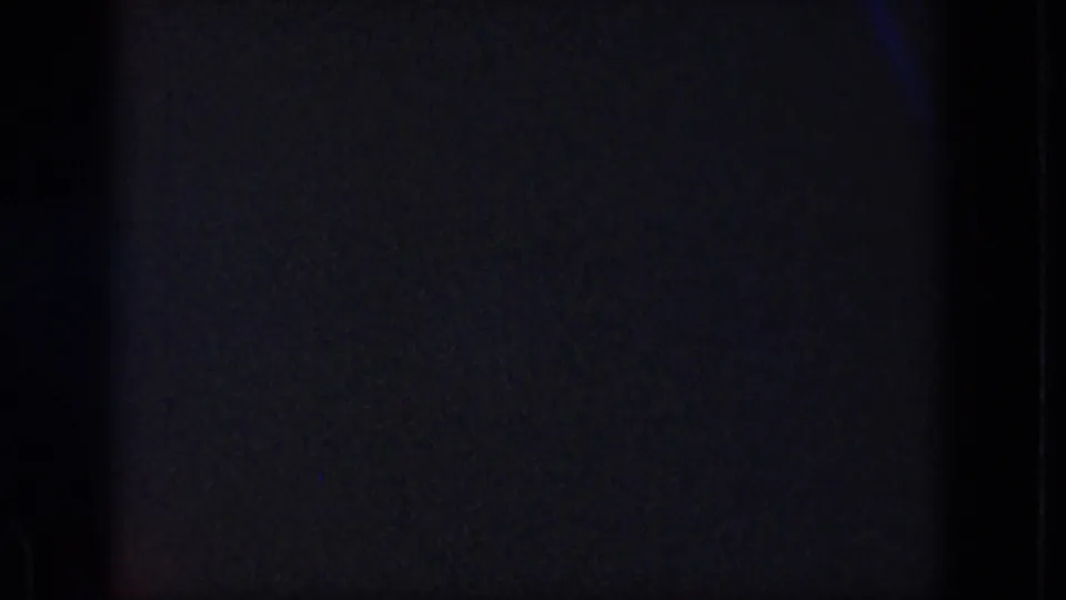 Low-exposure 8mm frame showing mostly dark film texture, vertical edge marks, and minimal scene detail during the clip opening.
