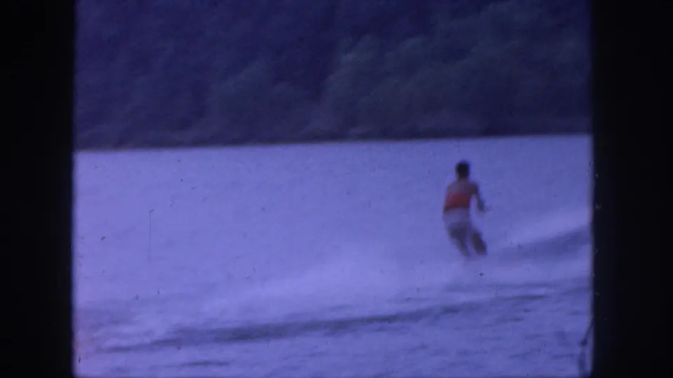 1961 Michigan home-movie frame showing a distant water skier on a lake, wake lines across the water, and a wooded shoreline behind them.