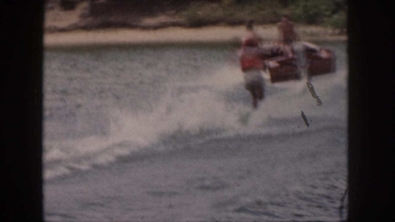 In 1961 Michigan home-movie footage, a water skier in an orange life vest follows a motorboat near a wooded shoreline.