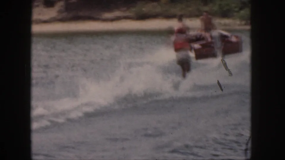 In 1961 Michigan home-movie footage, a water skier in an orange life vest follows a motorboat near a wooded shoreline.