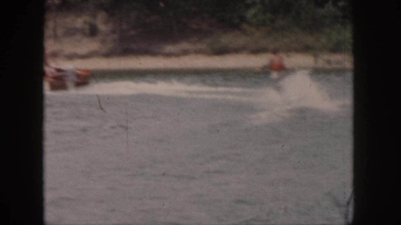 The skier appears farther from camera, trailing behind the boat across rippled water near a sandy wooded shore.