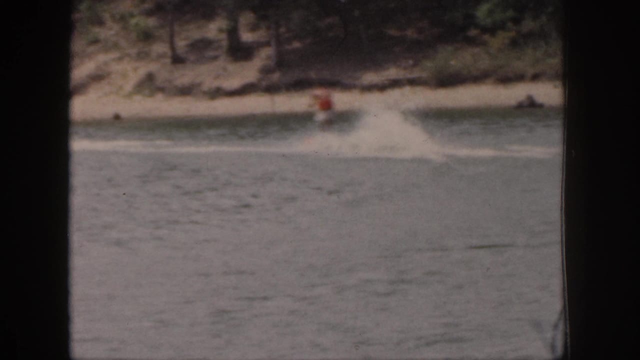 A distant skier in an orange vest crosses the lake wake near a wooded Michigan shoreline in faded 8mm color.