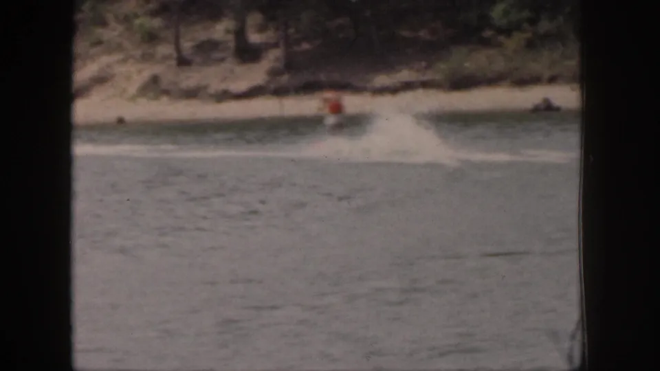 A distant skier in an orange vest crosses the lake wake near a wooded Michigan shoreline in faded 8mm color.