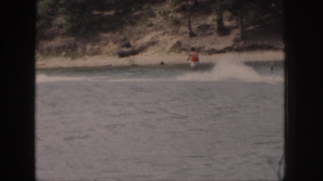 Distant water skier in a red top passes along a wooded lakeshore with spray behind him.