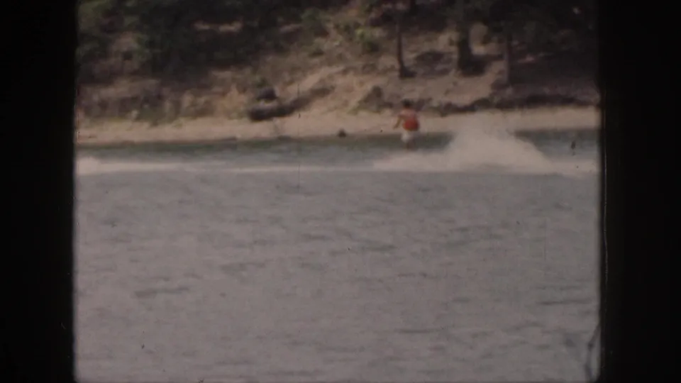 1961 Michigan 8mm frame showing a distant water skier near a sandy, tree-lined shore, seen through dark frame borders.