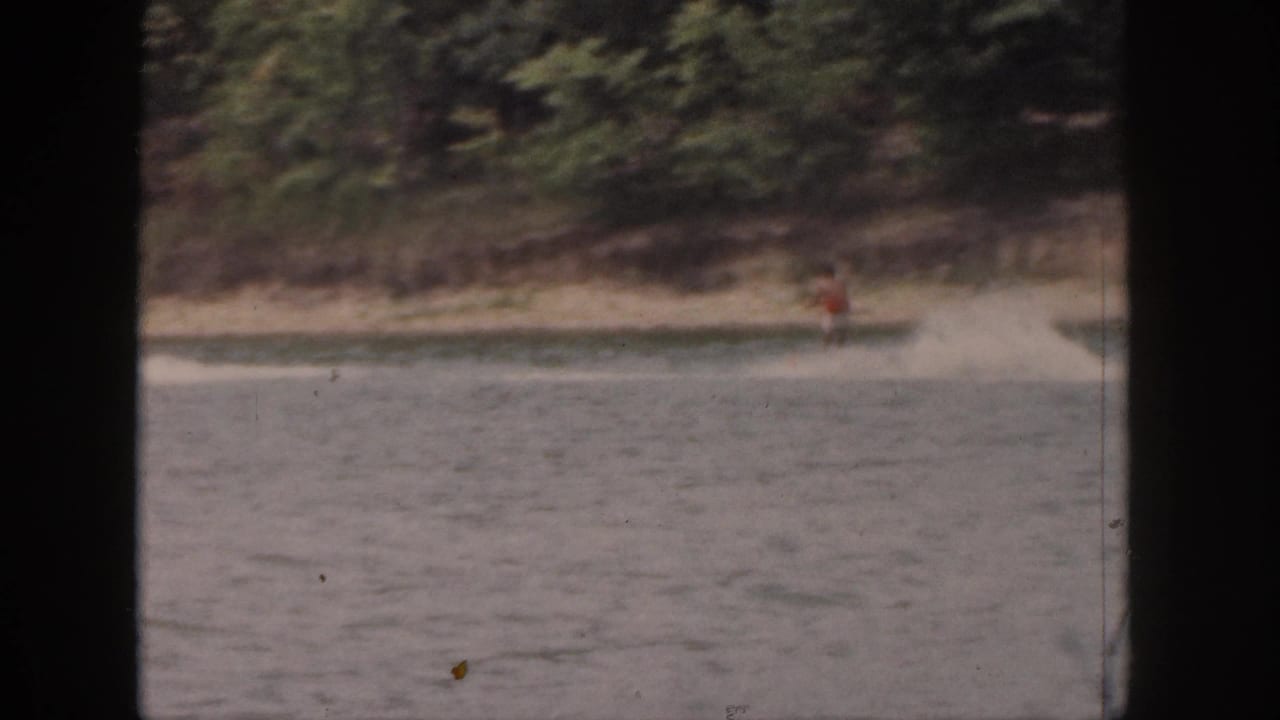 Water skier continues across the lake near dense trees and a pale shoreline.