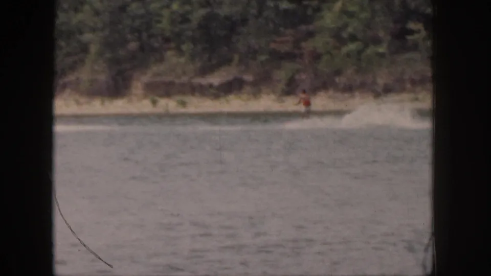 1961 Michigan home-movie frame with a distant water skier near a wooded shoreline and visible water spray.