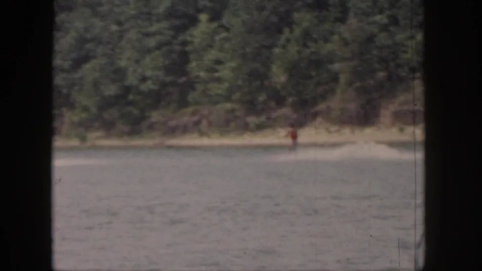 Soft 8mm frame from 1961 Michigan showing a distant skier near the right edge of a tree-lined lake shore.