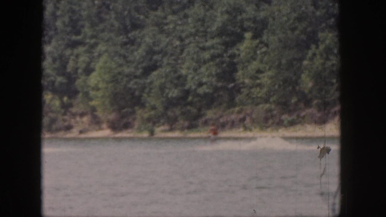 1961 Michigan 8mm frame showing a wooded lakeshore, calm lake water, and a distant person near the shoreline.