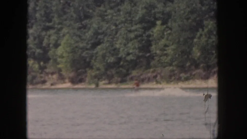 1961 Michigan 8mm frame showing a wooded lakeshore, calm lake water, and a distant person near the shoreline.