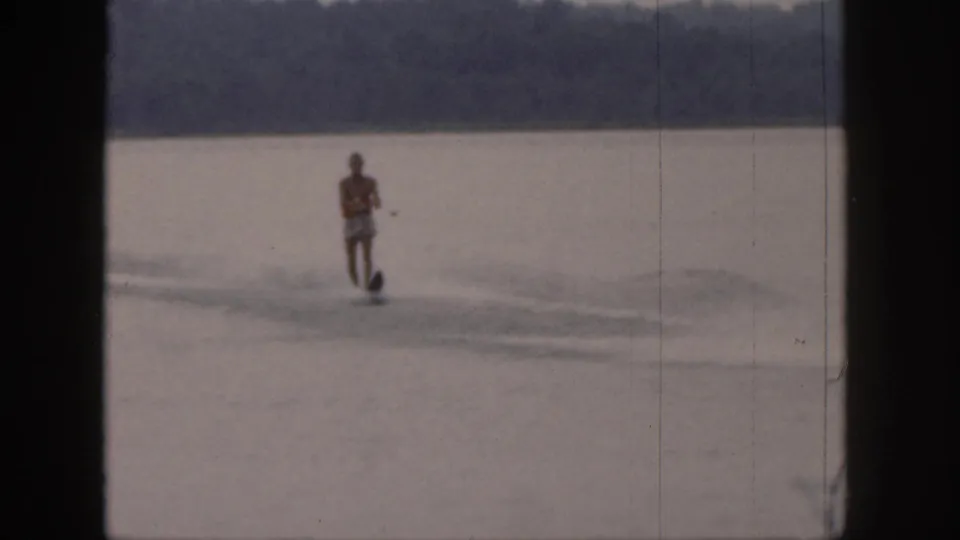 1961 Michigan 8mm frame of a water skier in a red lifejacket crossing calm lake water with a wooded shoreline beyond.