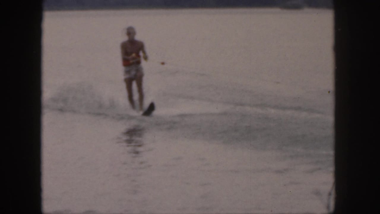 A shirtless man in patterned trunks water skis toward the camera on a Michigan lake, holding a tow rope.