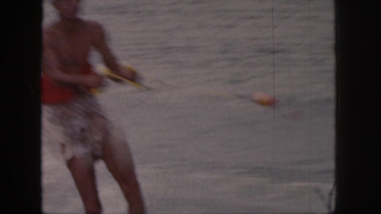 A close, motion-softened frame shows the shirtless skier gripping a yellow tow rope as he passes on the lake.