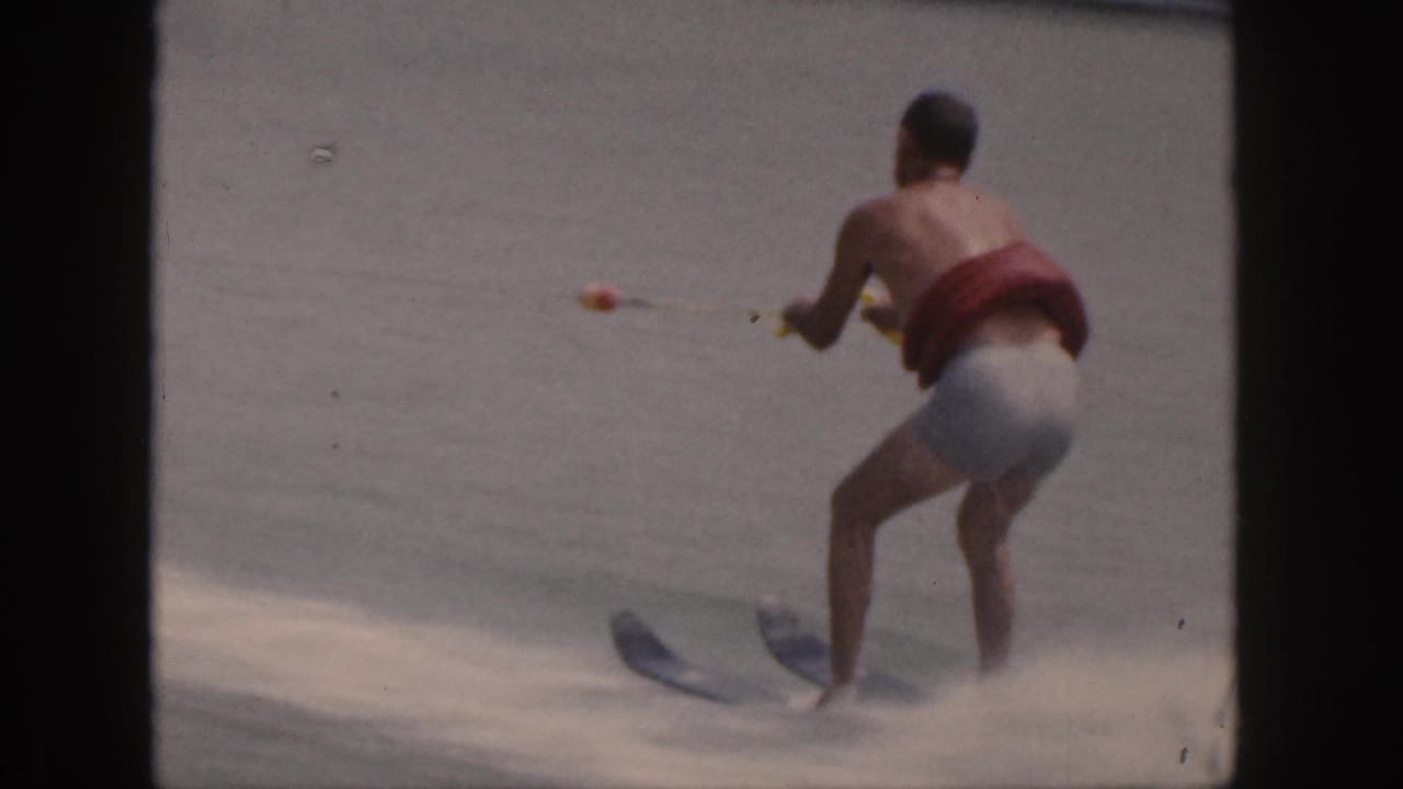 Shirtless man in a red life vest water skiing across a lake while holding tow ropes.